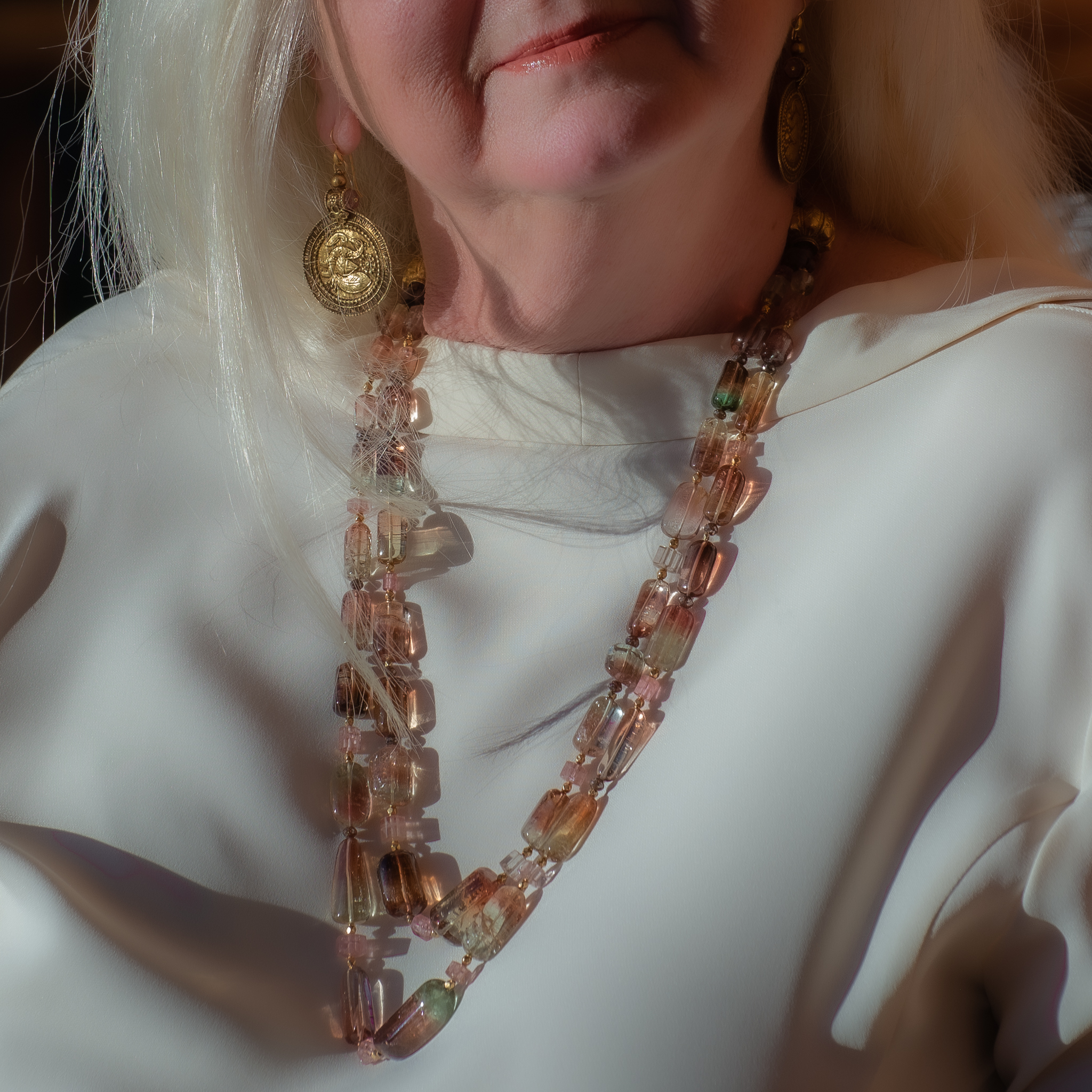 Photograph of the designer wearing gold medallion earrings with peacock motif and pink and green tourmaline crystal necklace